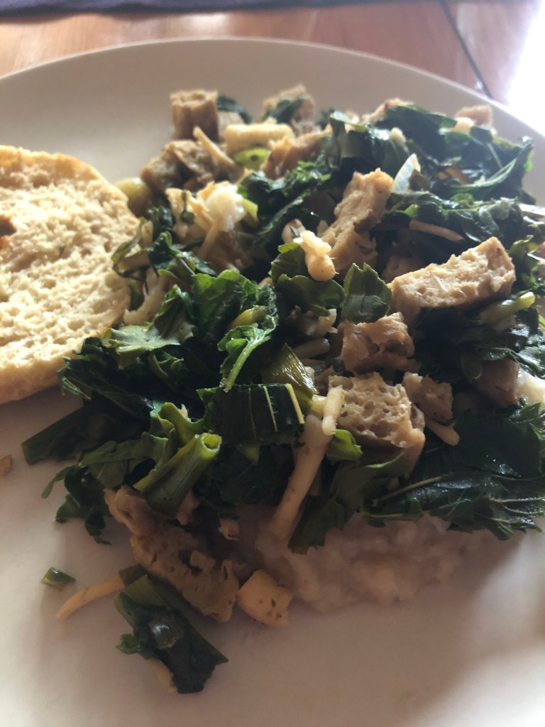 On a light tan plate, a half of a homemade bagel, mashed sunchokes with stir fried nettles, arugula, bell flower greens and bell flower roots, and seitan